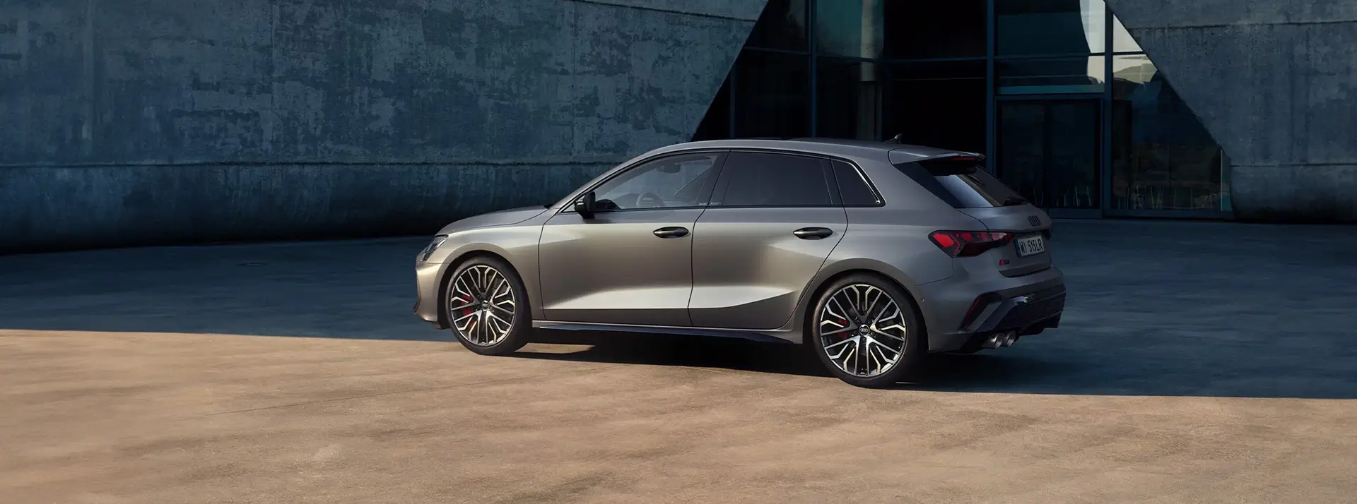 szare Audi S3 Sportback stoi w cieniu przed nowoczesnym betonowym budynkiem