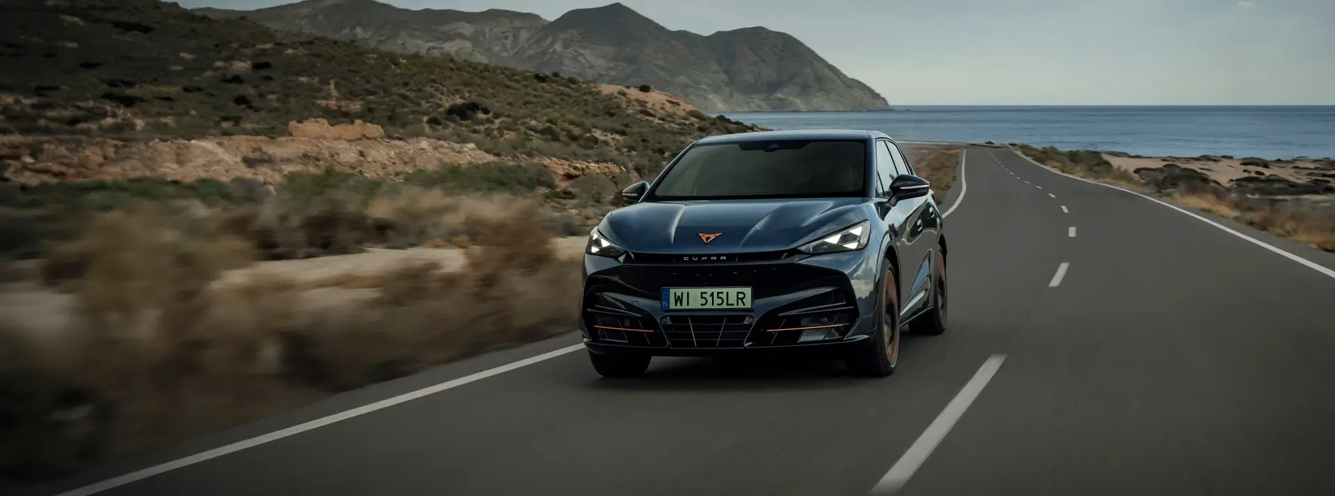 sportowy ciemnoniebieski SUV - CUPRA Tavasccan jedzie drogą położoną pomiędzy morzem a górami