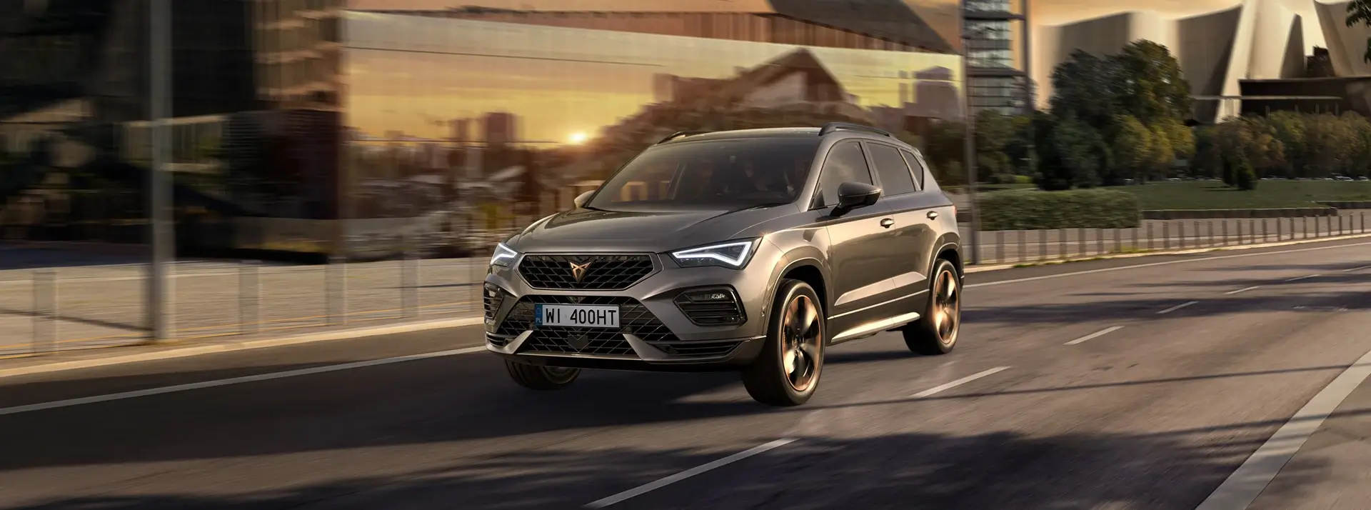 SUV CUPRA Ateca w szarografitowym kolorze jedzie drogą na tle rozmazanych budynków miasta